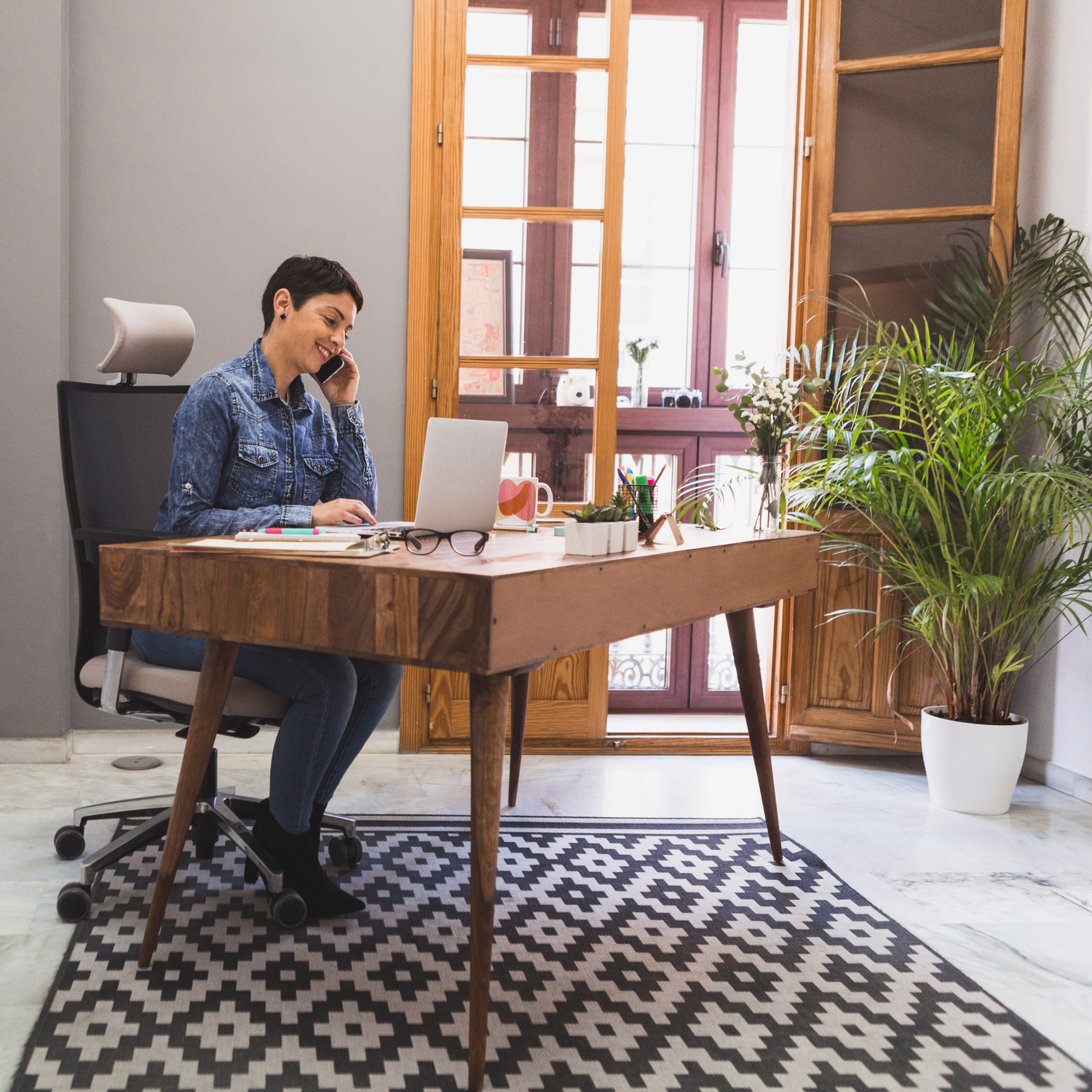 Een werkplek met een ergonomische bureaustoel en een houten tafel waar een vrouw aan het werk is op haar laptop.